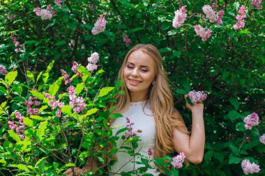 Leylak çalıyanında duran güzel beyaz elbise giyen büyüleyici sarışın bir kadın portresi.