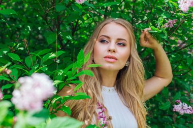 Leylak çalıyanında duran güzel beyaz elbise giyen büyüleyici sarışın bir kadın portresi.