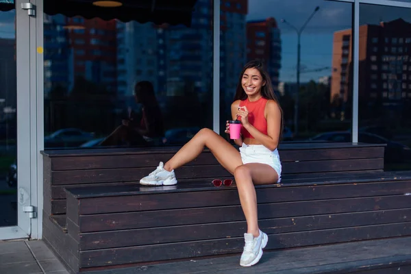 Elegante feliz jovem morena mulher vestindo branco shorts e tênis segurando rosa xícara de café para ir sentado ao lado de café . — Fotografia de Stock