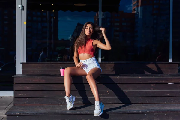 Stylish happy young brunette woman wearing white shorts and sneakers holding pink cup of coffee to go sitting next to coffee shop.