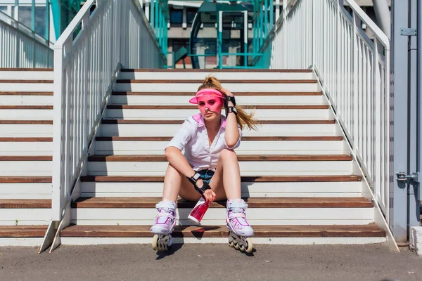 Potret seorang gadis emosional dengan topi pink mengenakan sarung tangan pelindung dan rollerblades duduk di tangga sambil memegang botol air di tangan — Stok Foto