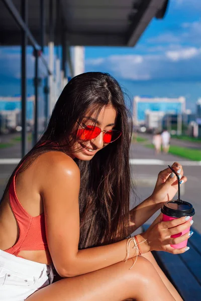 Elegante feliz jovem morena mulher vestindo branco shorts e tênis segurando rosa xícara de café para ir sentado ao lado de café . — Fotografia de Stock