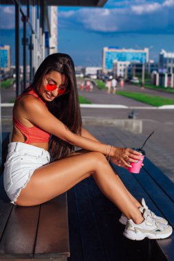 Şık mutlu genç esmer kadın beyaz şort ve spor ayakkabı kahve dükkanı yanında oturan gitmek için kahve pembe fincan tutan giyen.