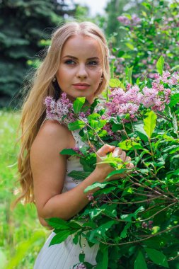 Leylak çalıyanında duran güzel beyaz elbise giyen büyüleyici sarışın bir kadın portresi.