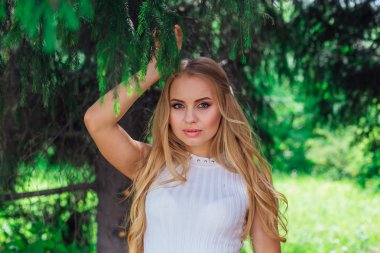 Kürk ağacının altında duran güzel beyaz elbise giyen büyüleyici sarışın bir kadın portresi.