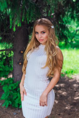 Kürk ağacının altında duran güzel beyaz elbise giyen büyüleyici sarışın bir kadın portresi.