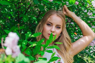 Leylak çalıyanında duran güzel beyaz elbise giyen büyüleyici sarışın bir kadın portresi.