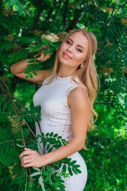 Rowan ağacının yanında duran güzel beyaz elbise giyen büyüleyici sarışın bir kadın portresi.