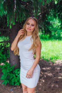 Kürk ağacının altında duran güzel beyaz elbise giyen büyüleyici sarışın bir kadın portresi.