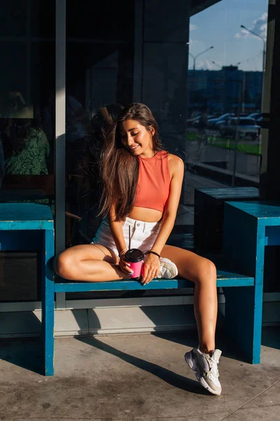 Stylish happy young brunette woman wearing white shorts and sneakers holding pink cup of coffee to go. — Stock Photo, Image