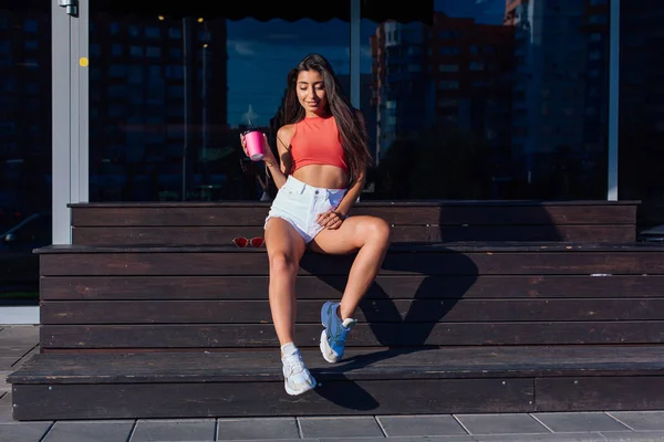 Elegante mujer morena feliz joven con pantalones cortos blancos y zapatillas de deporte con taza de café rosa para ir sentado al lado de la cafetería . —  Fotos de Stock