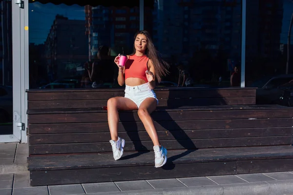 Elegante feliz jovem morena mulher vestindo branco shorts e tênis segurando rosa xícara de café para ir sentado ao lado de café . — Fotografia de Stock