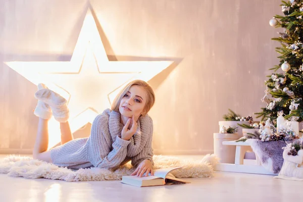 Mulher jovem bonita que põe e lê o livro abaixo da árvore de Natal em um interior de férias — Fotografia de Stock