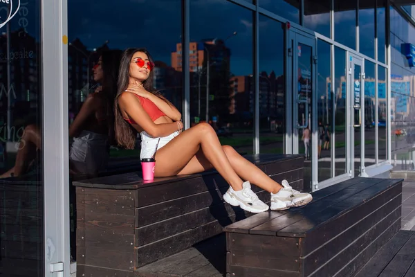 Elegante feliz jovem morena mulher vestindo branco shorts e tênis segurando rosa xícara de café para ir sentado ao lado de café . — Fotografia de Stock