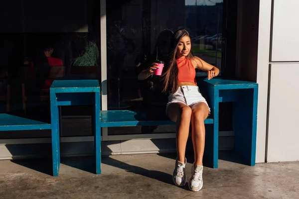 Snygg lycklig ung brunett kvinna klädd i vita shorts och sneakers håller rosa kopp kaffe att gå. — Stockfoto
