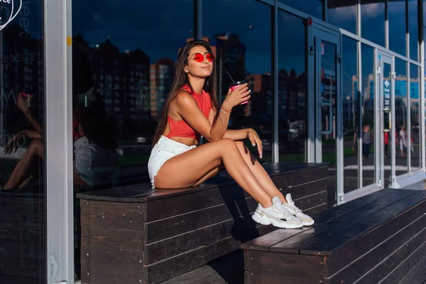 Elegante feliz jovem morena mulher vestindo branco shorts e tênis segurando rosa xícara de café para ir sentado ao lado de café . — Fotografia de Stock