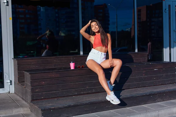 Elegante feliz jovem morena mulher vestindo branco shorts e tênis segurando rosa xícara de café para ir sentado ao lado de café . — Fotografia de Stock