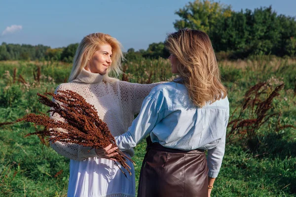 Portrait of two beautiful women in a field in late summer. Beautiful girls standing next to each other and holding bouquet made of dry brown plants. — Stock Photo, Image