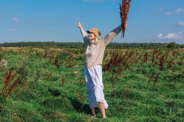 Mulher loira bonita feliz que anda em um campo verde com um bouquete de plantas marrons secas — Fotografia de Stock