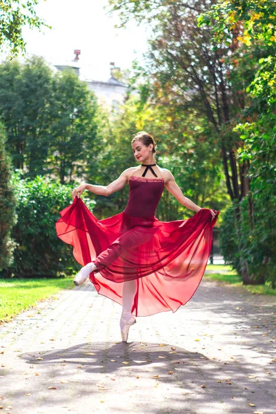 Woman ballerina in red ballet dress dancing in pointe shoes in autumn park.