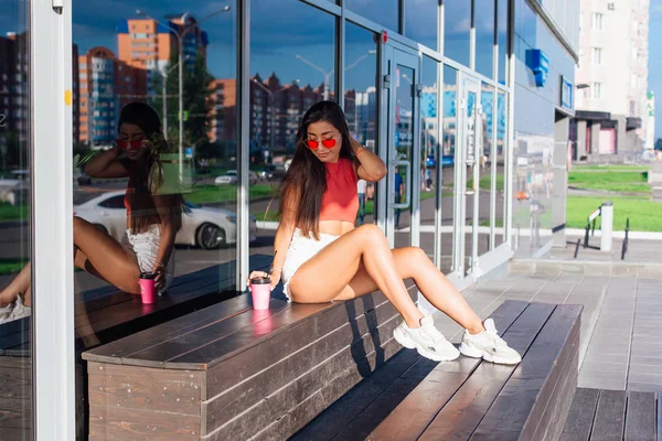Stylish happy young brunette woman wearing white shorts and sneakers holding pink cup of coffee to go sitting next to coffee shop. — Stock Photo, Image