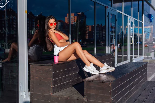 Snygg lycklig ung brunett kvinna klädd i vita shorts och sneakers håller rosa kopp kaffe att gå bredvid kafé. — Stockfoto