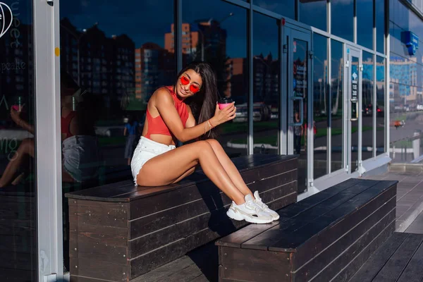 Elegante feliz jovem morena mulher vestindo branco shorts e tênis segurando rosa xícara de café para ir sentado ao lado de café . — Fotografia de Stock