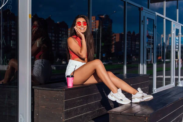 Snygg lycklig ung brunett kvinna klädd i vita shorts och sneakers håller rosa kopp kaffe att gå bredvid kafé. Stockfoto