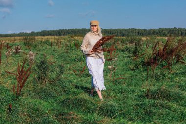 Yeşil bir tarlada bir buket kuru kahverengi bitkiyle yürüyen mutlu, güzel sarışın kadın.