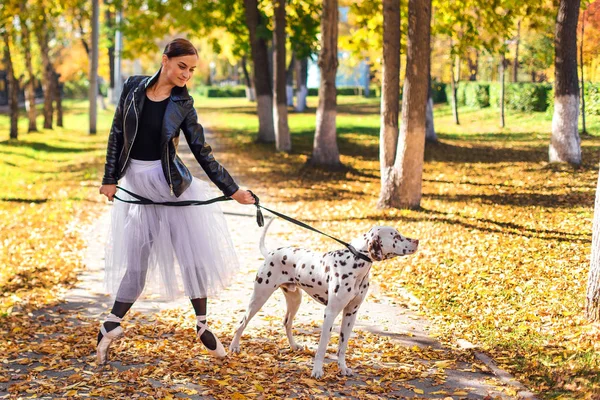Ballerina with Dalmatian dog in the Park. Woman ballerina in a white ballet skirt and black leather jacket dancing in pointe shoes in autumn park with her spotty dalmatian dog. — Stock Photo, Image