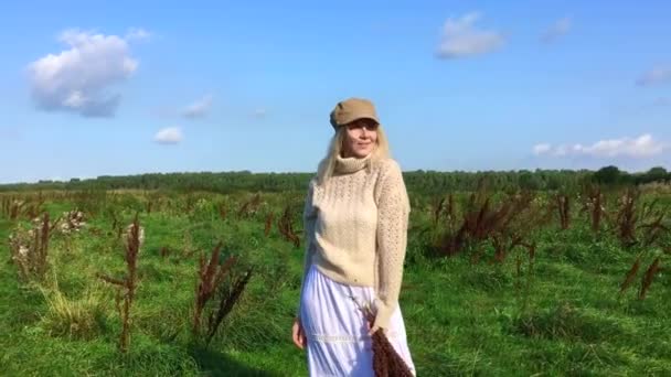 Yeşil bir tarlada bir buket kuru kahverengi bitkiyle yürüyen mutlu, güzel sarışın kadın. — Stok video