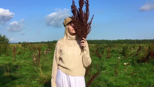 Mujer rubia hermosa feliz caminando en un campo verde con un ramo de plantas marrones secas — Vídeos de Stock