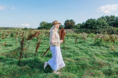 Yeşil bir tarlada bir buket kuru kahverengi bitkiyle yürüyen mutlu, güzel sarışın kadın.