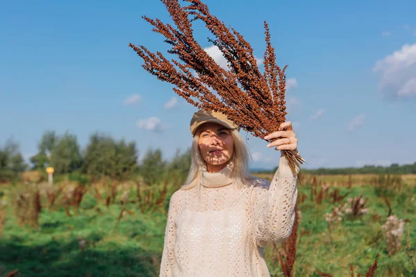 Mujer rubia hermosa feliz caminando en un campo verde con un ramo de plantas marrones secas — Foto de Stock