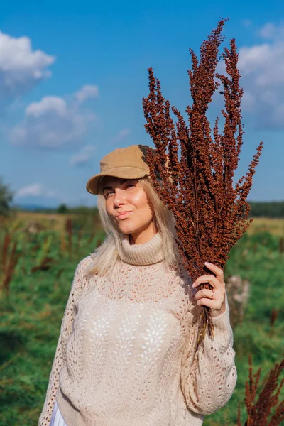 Happy beautiful blond woman walking in a green field with a bouquete of dry brown plants — Stock Photo, Image
