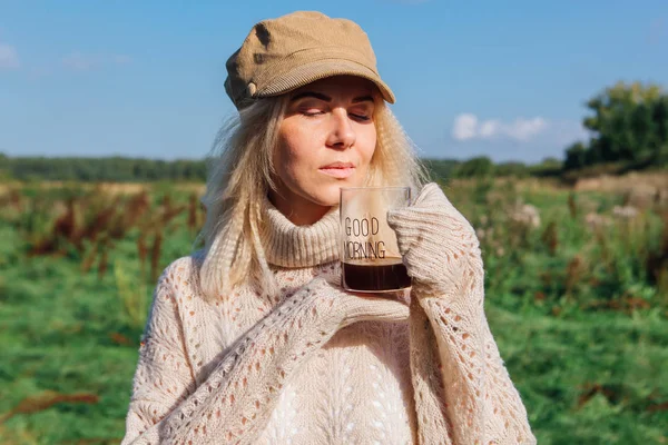 Mulher loira bonita segurando uma xícara de vidro de café em um campo — Fotografia de Stock
