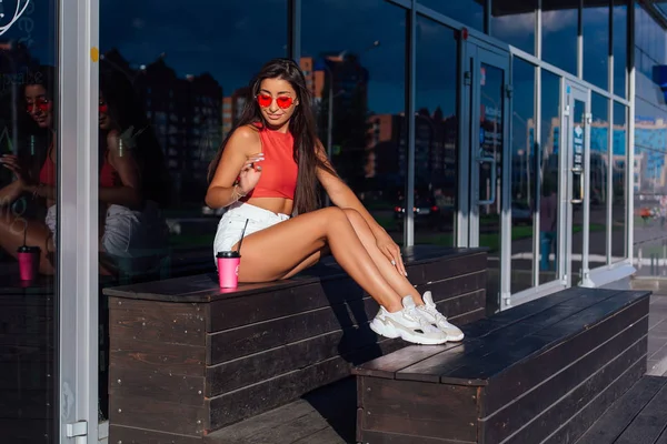 Elegante feliz jovem morena mulher vestindo branco shorts e tênis segurando rosa xícara de café para ir sentado ao lado de café . — Fotografia de Stock