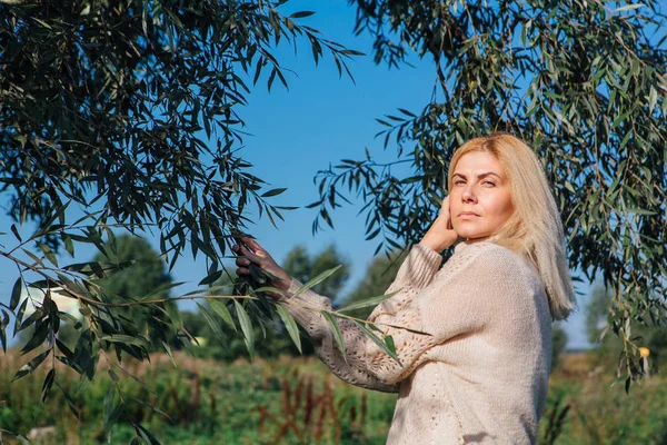 Ağacın yanında elinde yeşil yapraklı brunchlar tutan mutlu sarışın kadın.