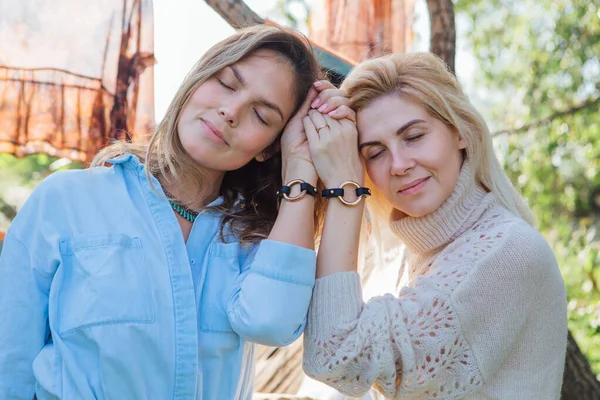 Portrait of two beautiful women brunette and blond holding their hands with same bracelets next to the tree in late summer. Beautiful girls standing next to each other. — Stock Photo, Image