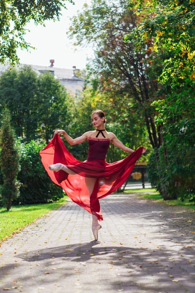 Woman ballerina in red ballet dress dancing in pointe shoes in autumn park.