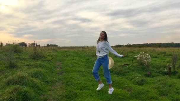 Happy beautiful brunette teenage girl walking on the road in a green field during beauriful sunset with colorful sky. Девушка бросает шляпу в небо — стоковое видео