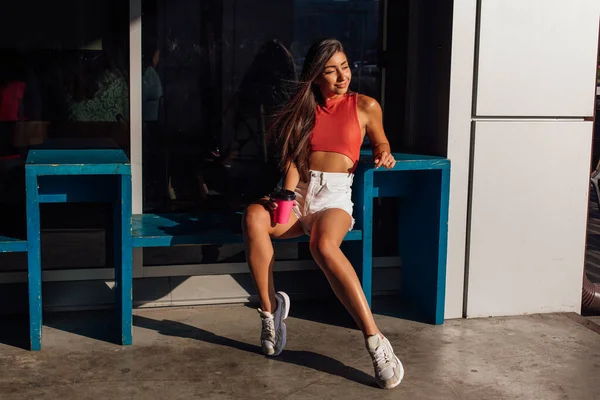 Elegante mujer morena feliz joven con pantalones cortos blancos y zapatillas de deporte con taza de café rosa para llevar . — Foto de Stock