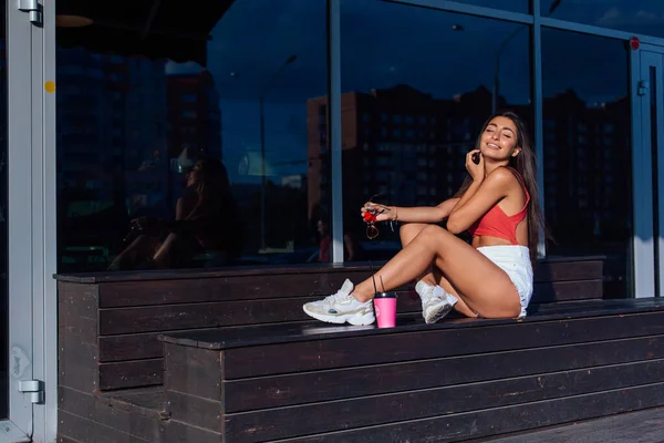 Elegante feliz jovem morena mulher vestindo branco shorts e tênis segurando rosa xícara de café para ir sentado ao lado de café . — Fotografia de Stock