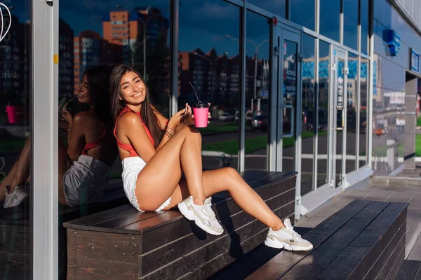Elegante feliz jovem morena mulher vestindo branco shorts e tênis segurando rosa xícara de café para ir sentado ao lado de café . — Fotografia de Stock