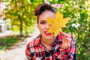 Makyajlı ve tokalı güzel bir kadın sokakta dikilmiş, yüzüne yakın sarı akçaağaç yaprağı tutuyor..