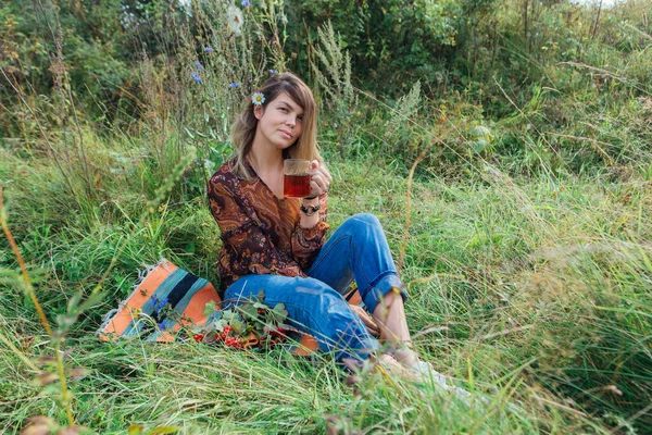 Bella donna con fiore di camomilla in capelli seduti sull'erba e bere tè all'aperto — Foto Stock