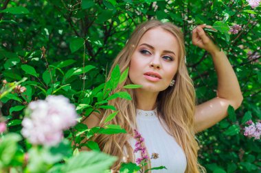 Leylak çalıyanında duran güzel beyaz elbise giyen büyüleyici sarışın bir kadın portresi.