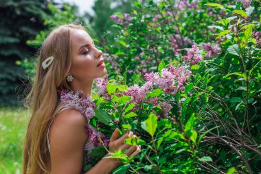 Leylak çalıyanında duran güzel beyaz elbise giyen büyüleyici sarışın bir kadın portresi.