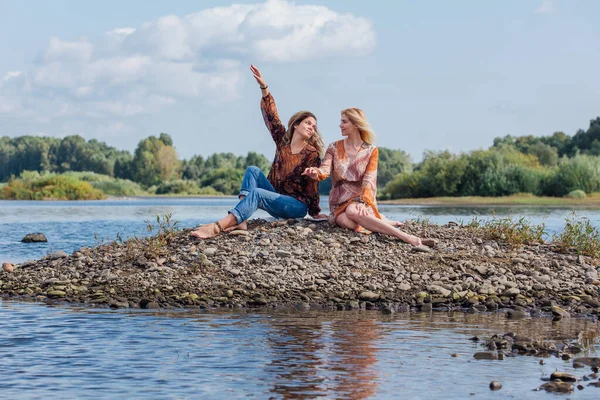 Two pretty sisters sitting on the small stone river island on a cloudy windy summer day, discuss girls secrets, laugh. — Stock Photo, Image