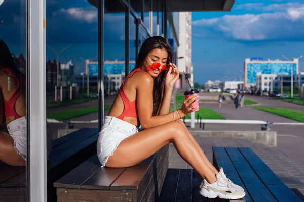 Elegante feliz jovem morena mulher vestindo branco shorts e tênis segurando rosa xícara de café para ir sentado ao lado de café . — Fotografia de Stock